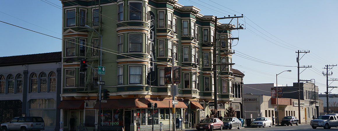 A multi-story green and beige building with bay windows and awnings, situated on a street corner with cars parked along the side.